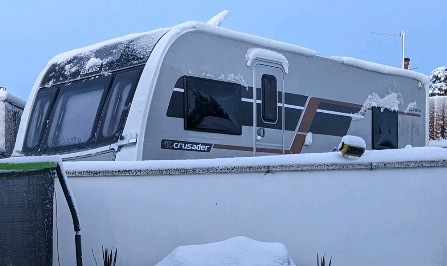 A snow covered Elddis Crusader Aurora caravan parked behind a wall in winter